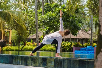 Selbstklebende Fototapeten Entspannung Beautiful young woman is practicing yoga on the edge of swimming pool with green plants and palm trees around. Sport photo with beautiful girl in luxury hotel on Bali island Indonesia  © Alexander