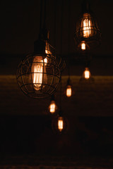 Fototapeta na wymiar Industrial style lanterns hanged from ceiling. Metal wire lampshades in darkness. Lamps with glowing filaments inside Edison glass light bulbs. Urban style interior lighting. Wire cage lamps closeup