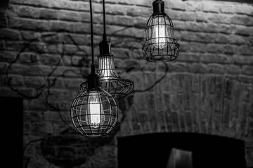 Black and white photography of wire lamps hanged from ceiling on old brick wall background. Glowing...