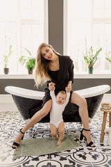 young beautiful blonde girl in stylish classic clothes of black color sits in a bathroom with a baby in a white apartment