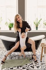 young beautiful blonde girl in stylish classic clothes of black color sits in a bathroom with a baby in a white apartment