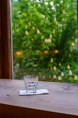 One glass of water stands on a white napkin on a wooden table near a window overlooking the avenue with blooming chestnut trees. Art photography