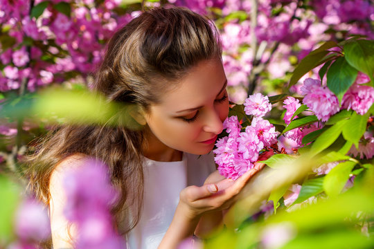 Beautiful young girl sniffs pink flowers, blooming sakura in spring garden. Beauty fashion photo shoot