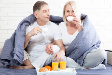 Portrait of happy playful couple relaxing in comfortable cozy bed looking at each other, cheerful man and woman having fun posing. Holding cups of coffee while covering their shoulders with blanket.