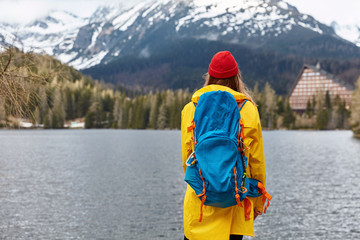Outdoor shot of female hiker stands back at camera with big blue rucksack, wears yellow raincoat and red headgear, poses at coast of mountain lake, admires majectic nature scenery. Snow mountains