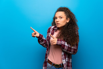 Teenager girl over blue wall frightened and pointing to the side