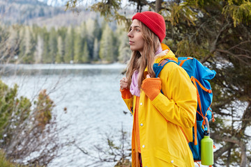 Sideways shot of beautiful female traveler stands with backpack, focused into distance pensively, wears red headgear and yellow coat, poses near picturesque lake and green trees with copy space