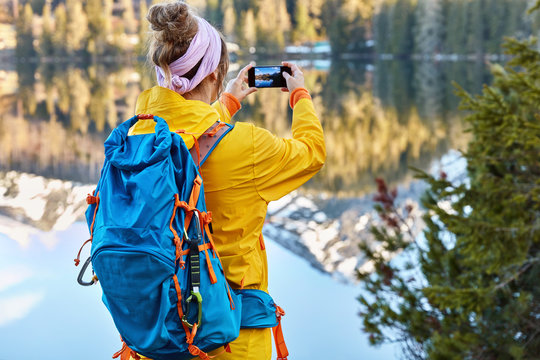 Woman traveler carries backpack takes phoro of beautiful lake landscape on smart phone, enjoys nature, mountains reflect in water, green fir grows near. Tourism, vacation and travel concept.