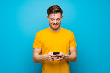 Redhead man over blue wall sending a message with the mobile