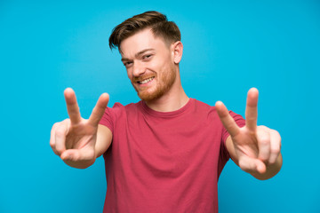 Redhead man on isolated blue wall smiling and showing victory sign