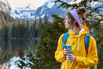 Naklejka premium Pensive female mountaineer wears scarf around head, yellow raincoat, enjoys hot coffee from flask, has trip in mountains, poses on nature landscape with copy space, hikes in beautiful forest