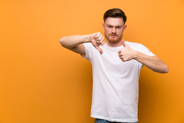 Redhead man over brown wall making good-bad sign. Undecided between yes or not