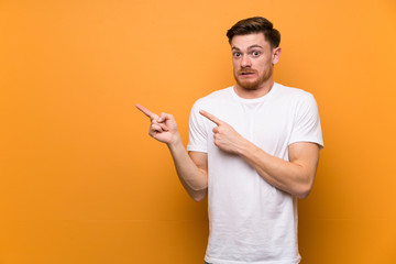 Redhead man over brown wall frightened and pointing to the side