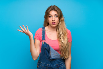 Fototapeta premium Young blonde woman with overalls over isolated blue wall making doubts gesture