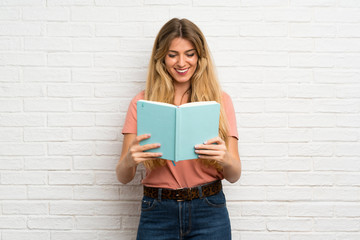 Young blonde woman over white brick wall holding and reading a book