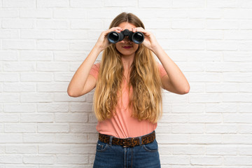 Young blonde woman over white brick wall with black binoculars