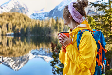Naklejka premium Pensive woman watches scenery, drinks coffee from disposable cup, wears yellow raincoat, carries backpack, has coffee break, likes being alone with nature, leads active lifestyle. People, recreation