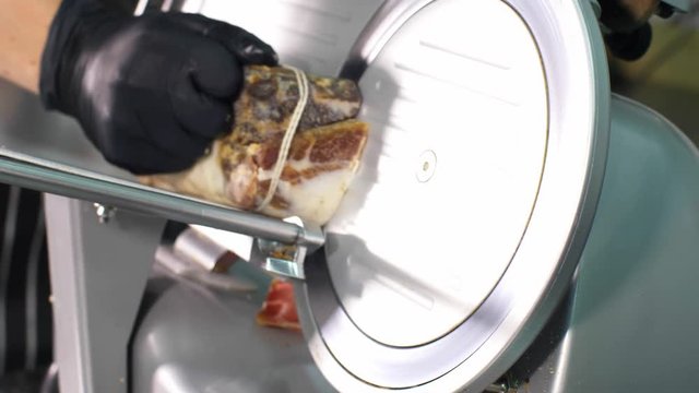 Man in black gloves is slicing a piece of smoked meat, moving a piece of meat to metal circle disc blade. Chef using automatic slicer machine, hand closeup.