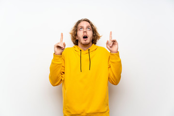 Blonde man with  sweatshirt over white wall surprised and pointing up