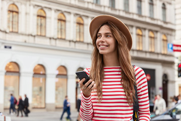 Fototapeta premium Happy millennial hipster girl wears stylish headgear and striped jumper, holds modern cellular for communication, enjoys walk in city, receives good message, searches for showplace or route online