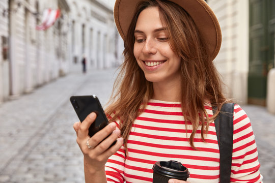 Cropped image of charming tourist uses application on cellular, searches direction for walking in sightseeing tour, enjoys drinking hot beverage wears stylish hat and striped jumper. Traveling concept