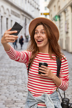 Overjoyed European Female Traveler In Hat, Makes Selfie Portrait Outdoor, Has Fun During Excursion In Ancient Town, Drinks Coffee From Disposable Cup, Wears Stylish Hat, Laughs Joyfully At Camera