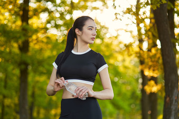 Sexy, sporty girl with a beautiful figure wearing a sports brassiere and leggings,stands with a bottle of water in her hands in the background of the nature landscape on the rest from sports exercises