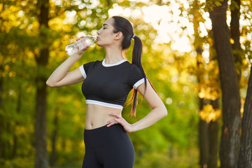 Sexy, sporty girl with a beautiful figure wearing a sports brassiere and leggings,stands with a bottle of water in her hands in the background of the nature landscape on the rest from sports exercises