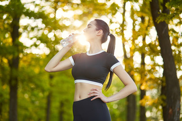 Sexy, sporty girl with a beautiful figure wearing a sports brassiere and leggings,stands with a bottle of water in her hands in the background of the nature landscape on the rest from sports exercises