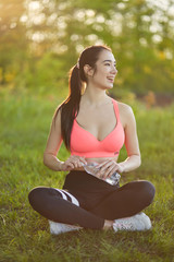 Sexy, sporty girl with a beautiful figure wearing a sports brassiere and leggings, sitting with a bottle of water in her hands in the background of the nature landscape 