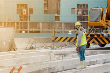 The construction worker in the green waistcoat and the yellow helmet and a black mask standing  on the steel beam in the construction area with yellow crane in the background.