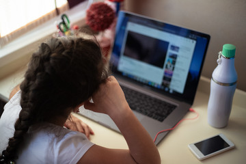 Little girl looking in laptop on his homework table near thermo bottle and other supplies