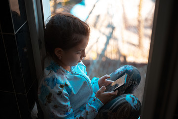 Portrait of modern children girl sitting on the windowsill with smartphone in hands