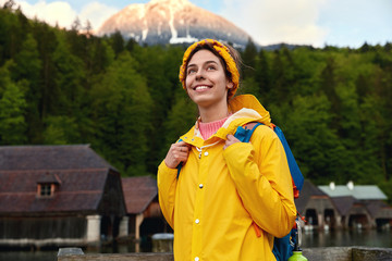 Half length shot of happy carefree woman tourist in yellow active wear, spends time away from home, recreats in nice mountain place, lives in small house near green forest, blue sky in background