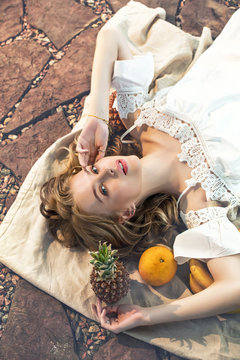 Young beautiful happy blonde model girl lying on a linen bedspread with fruit