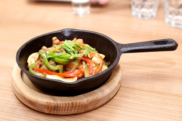 A small cast-iron skillet with food with red pepper and green onions standing on wooden sub-base on wooden table