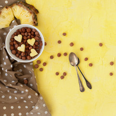Breakfast of cornflakes, milk and fruit, pineapple on a light yellow background. Pieces of pineapples in the form of hearts.Top view.