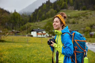 Horizontal view of cheerful hitch hiker explores beautiful scenic nature places, walks across green hill near mountains, makes photo of small houses over majestic landscape, wears casual clothes