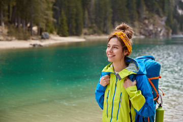 Outside shot of cheerful backpacker in high spirit, walks near turquoise lake in mountains, green trees, enjoys magical view, blank copy space area aside for text, visits popular tourist attraction