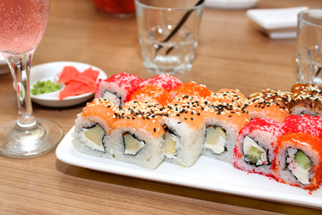 Sushi with fish, cheese and caviar on a white rectangular plate, a glass of pink soda, empty glasses in the background