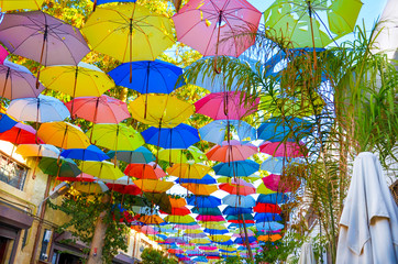 Colorful umbrellas decorating the top of the street in Cypriot Nicosia. The umbrella serves also as a shade and protection against the sunshine. Among the umbrellas there are green tree branches - obrazy, fototapety, plakaty