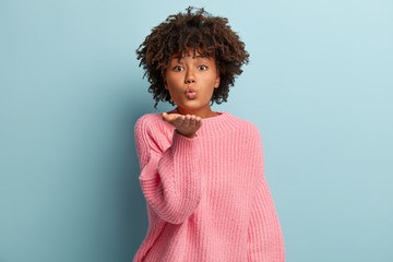Glamorous dark skinned woman sends air kiss, keeps lips folded, has Afro haircut, wears casual pink jumper, isolated over blue background, says be valentine. Flirting and body language concept