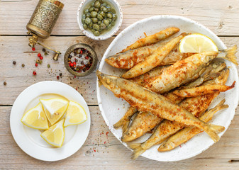 Fried smelt in a white plate. Small fish. Capers, lemon, pepper and salt on a wooden table. A delicious dinner in the rustic style. Selective focus