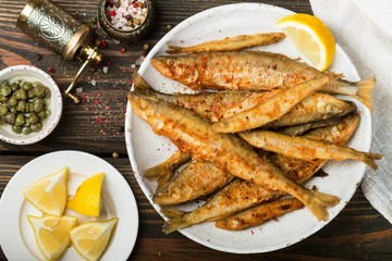 Fried smelt in a white plate. Small fish. Capers, lemon, pepper and salt on a wooden table. A...