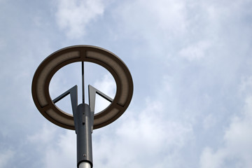 A grey round lamp with an led base with black three branched holders mounted on a cylindrical lamp post against a blue sky covered with white clouds