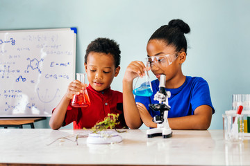 Cheerful black pretty smart children in casual clothes sitting in school chemistry laboratory and holding flasks with colourful liquids