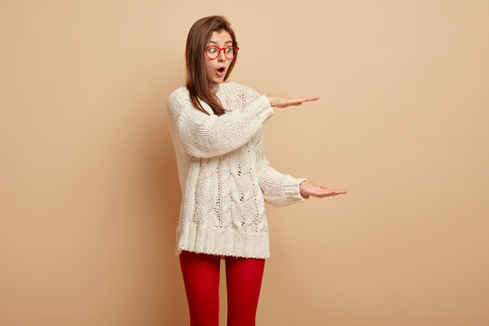 Half length shot of astonished shocked lady describes size of something big with surprised look, shapes huge item against brown background, wears casual jumper and leggings. Body language concept