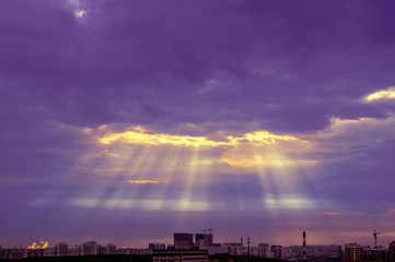 Sun rays through an opening in the cloudy purple sky. Violet landscape with yellow sun lights in the city on horizon
