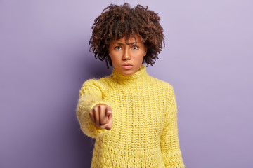 Displeased woman has Afro hairstyle, blames someone, points directly at camera, makes serious face, wears winter yellow sweater, stands against purple background. You are guilty in my misfortune
