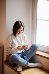 Young woman watches something on the Internet in her mobile phone and smiling while sitting on a windowsill